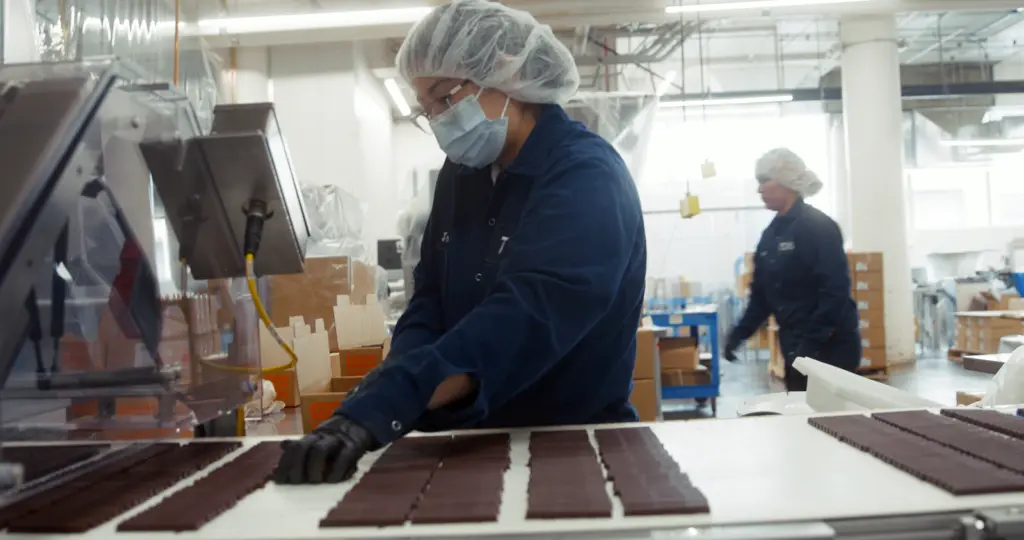 Food processing worker in protective equipment handling chocolate on production line.