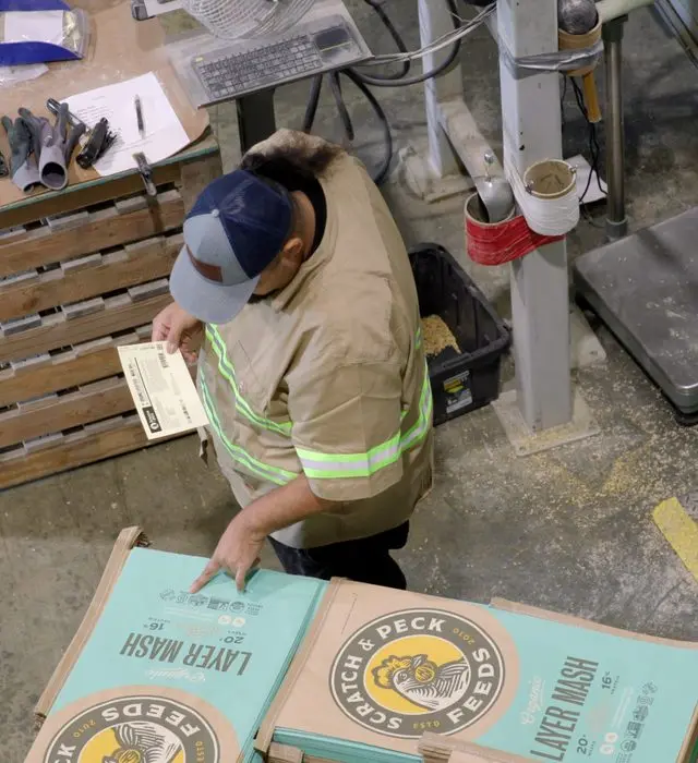 Overhead view of a worker in a hi-vis jacket and ball cap on a mill floor, holding a printed tag in one hand and pointing at the label panel of a stack of Scratch and Peck organic layer mash feed bags.