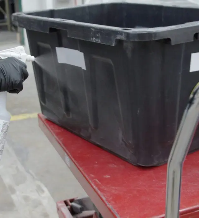 A gloved hand sprays cleaning solution from a trigger bottle onto a black plastic tote resting on a red scissor-lift cart in a processing area.