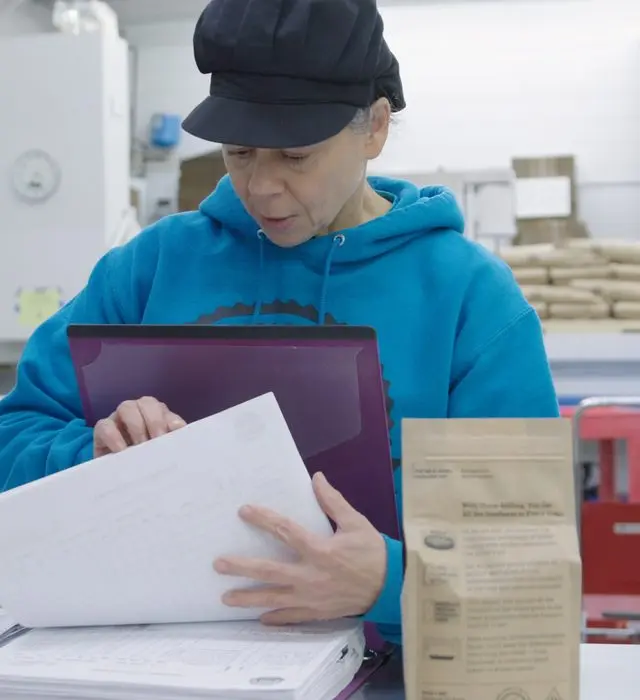 A woman in a black work cap and blue hooded sweatshirt reads a printed record sheet from a binder at a mill workbench, a retail bag of stone-milled flour beside her and sacks stacked on a pallet behind.