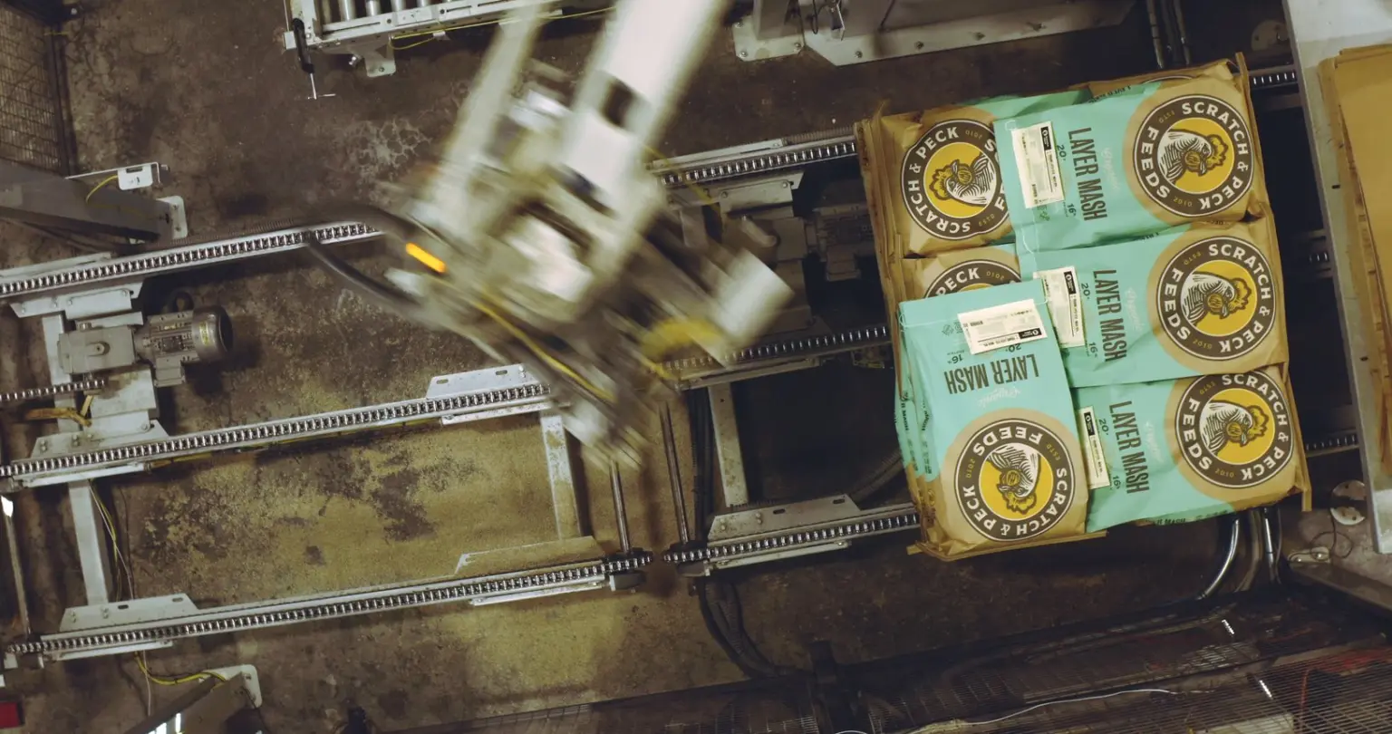 Overhead view of a robotic claw moving Scratch & Peck feed bags beside stacked bags in a packing area