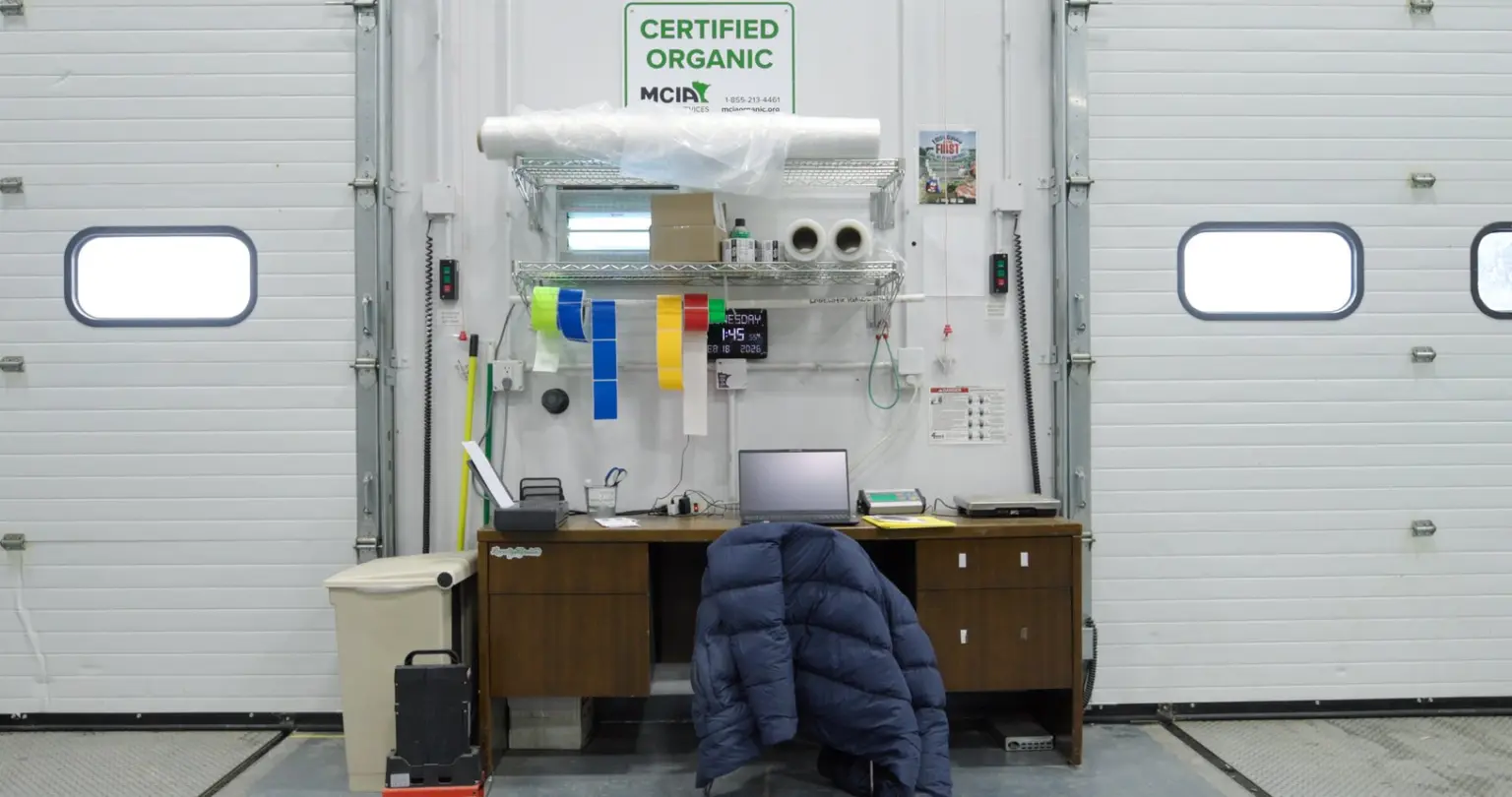 Compliance work station at a facility door with rolls of colored labeling tape, a laptop and a scale beneath a Certified Organic sign