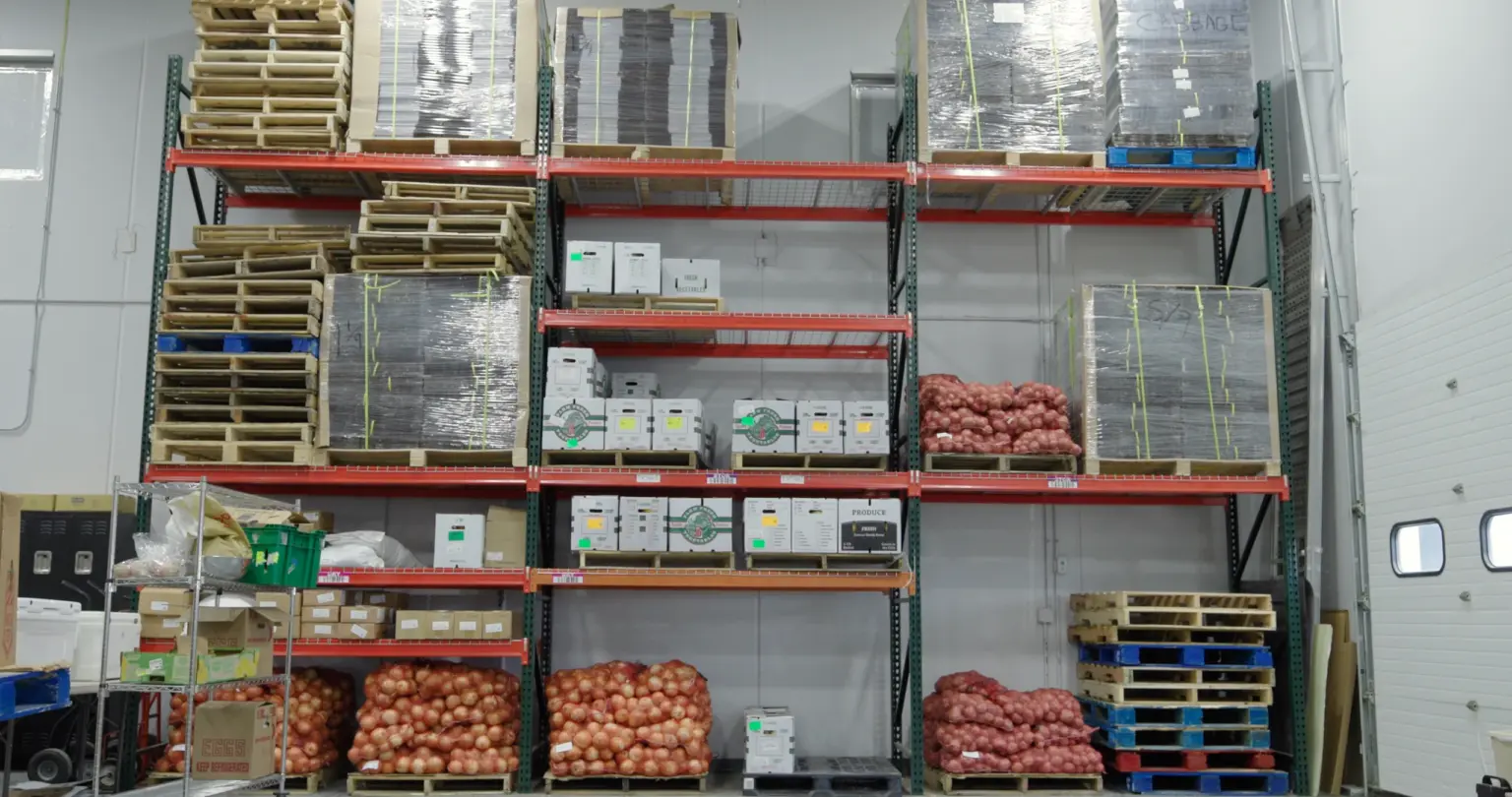 Pallet racking stacked with boxed produce and sacks of onions in an organic warehouse.