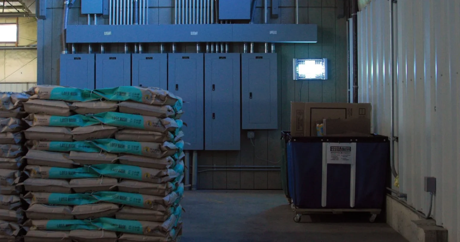Illuminated insect light trap mounted on a wall above stacked feed bags in a warehouse, beside electrical panels and a recycling bin